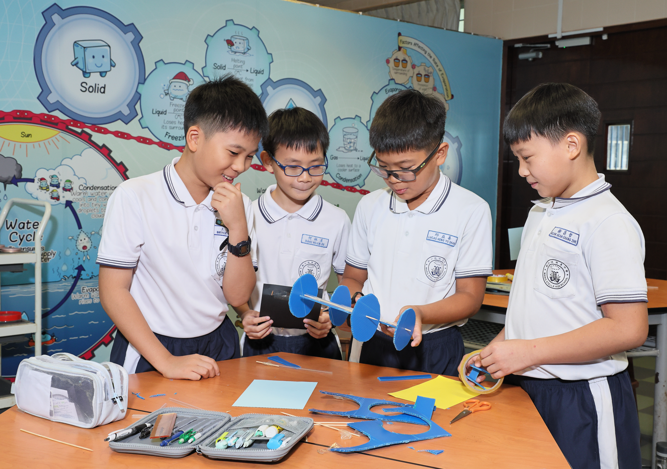 Four masked schoolboys in white uniforms conduct a hands-on science experiment at a classroom table.